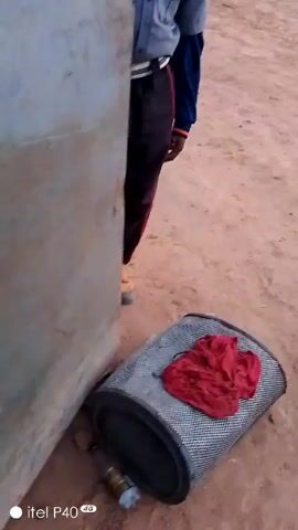 The video shows a person wearing a blue shirt and dark pants standing outside, partially obscured by a wall. There is a stool with a red cloth on it in the foreground. The camera then moves to show a dirt area with small buildings in the background. The camera moves again, focusing on what appears to be the interior of a building with a wall and a partially visible window or door. The camera movement is shaky and the focus is inconsistent.