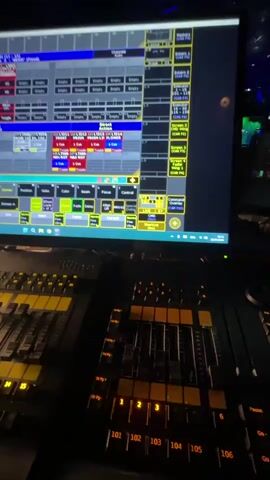 The video shows a close-up of a computer screen displaying a complex lighting control interface with multiple rows of buttons and sliders. Below the screen, a portion of a physical lighting control console with numerous faders is visible. The scene is dimly lit, with the screen and console illuminated by their own lights. In the background, a nightclub or similar venue can be seen with people moving around and colorful, flashing lights, primarily green and blue, creating a vibrant atmosphere.