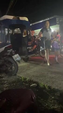 The video shows a street scene at night with several people and vehicles moving around. A blue and white tricycle with the number 654 on the back is parked. Beside it is a white motorcycle with a license plate that reads "L641KD". People walk past the tricycle and motorcycles drive past. The light source appears to be overhead streetlights. The ground is a paved surface with grass and small rocks visible on the side. In the foreground, a burgundy plastic stool is partially visible. A foot appears briefly in the bottom left corner of the frame. The overall scene appears to be a normal night at a roadside area.
