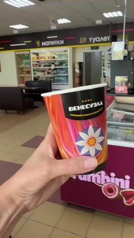 A hand holds a paper coffee cup with a colorful design and Russian text that reads "КОФЕ ВЕНЕСУЭЛА" and "Глубокий и многогранный вкус". The cup is positioned in the foreground. The background shows the interior of a convenience store. In the mid-ground, there are several refrigerated display cases, one with a sign that says "frambini" and displays frozen products. Behind the display cases, shelves are stocked with various items, including drinks and packaged goods. A couch and tables are visible further in the background, along with a checkout counter and overhead lighting. The overall environment appears to be a brightly lit retail space.