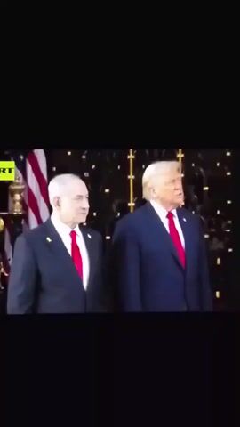 Two men stand side-by-side, both wearing dark suits and red ties. The man on the left has white hair and is looking to his right. The man on the right has blond hair and is speaking. Behind them is an American flag and a decorative backdrop with lighting. A logo that reads "RT" is visible in the top left corner of the screen.