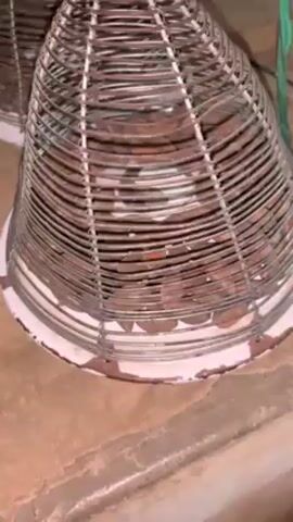 The video shows two percussion instruments, perhaps shakers, sitting on the floor. They have conical wire frames with decorative red cord at the top and a white base, and are filled with small brown objects, possibly seeds or beans. A green wire is partially visible near one of the instruments. The floor is a light brown color. A drum beat and shaker sound are heard in the background. The camera angle is close and slightly angled down.