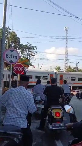 A train crosses a railway track at a railway crossing. A group of motorcyclists are waiting at the crossing, stopped by a red light and stop sign. There is a sign indicating a maximum height of 6.1 meters. The sky is clear and blue, with a cell tower visible in the background.