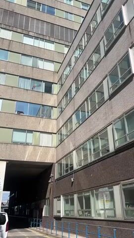 The video shows an upward shot of a tall, concrete building under a clear blue sky. The building has many rows of windows. To the right of the main building is a smaller building with brick on the lower level and a concrete facade with many windows on the upper levels. The camera pans upward, emphasizing the height of the main building. A dark passageway runs underneath the buildings. A white van is parked near the entrance to the passageway.