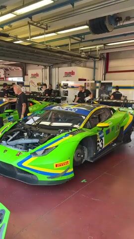 The video shows a race car garage. Several people are working on bright green racing cars, with their hoods open. One person is holding a helmet. Another scene shows a young man walking on a track outside the garage, holding a water bottle. The video appears to be shot from a handheld camera.