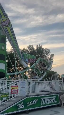 A pendulum amusement ride with a green and white color scheme swings people against a cloudy sky. The ride is labeled "3G 360 Turbo." Trees are visible in the background. The ride is at an amusement park, evidenced by the fencing and signage. The ride appears to be in motion, with passengers seated and secured.