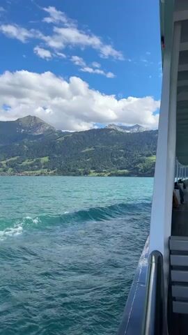 A video shows a view from a boat on a turquoise lake. The boat is moving, as evidenced by the waves and the wake behind it. Mountains covered in trees rise from the opposite shore, and some snow-capped peaks are visible in the distance. The sky is blue with scattered white clouds. The right side of the frame shows the railing and seating area of the boat.