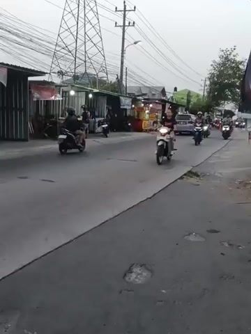 A video shows a street scene with several people riding motorcycles. The street is paved and has some potholes. There are buildings and stalls on the side of the road. There are also power lines and a large tower in the background. The sky is overcast. In the foreground, a person is riding a white scooter. In the middle ground, several other people are riding motorcycles. In the background, more motorcycles and a truck are visible. One person on a motorcycle appears to be carrying a child.