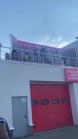 On a raised platform with a large pink banner in the background reading "RUSSIAN DRIFT SERIES RDS GP," three individuals in racing attire are celebrating. They are spraying champagne from bottles, creating a mist of liquid that falls around them. Two of the individuals are spraying the third person who is bending over. To the left of the group, a person wearing a bright yellow vest, possibly a photographer or official, is observing the celebration. Below the platform, a large red garage door is visible, with the number "2" appearing on a white sign to the left of the door. The sky above is overcast.