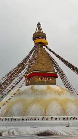 The video shows the Boudhanath Stupa in Nepal. It begins with a close-up view of the top of the stupa, adorned with colorful prayer flags. The camera then pans out to show the surrounding area, revealing a bustling marketplace with numerous people. The camera then focuses on the architecture of the stupa itself. The video concludes by going back up and again focusing on the top of the stupa.
