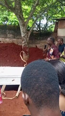 The video shows a burial service taking place outside, near a tree and a low wall. A white coffin is visible in the ground, presumably about to be buried. A man in a patterned shirt speaks into a microphone while holding a piece of paper. Several people are standing around, facing the coffin. The ground around the grave is reddish-brown soil. The overall tone seems somber.