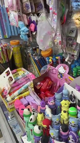 The video shows a brightly lit store selling stationery and accessories. The camera pans across shelves stocked with colorful items like pencils, pens, erasers, hair accessories, and small toys. In the background, a person wearing a dark shirt and pants can be seen browsing the merchandise. The store appears to be a small retail establishment with a variety of items displayed.