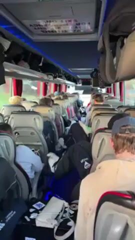 The inside of a bus filled with passengers is captured in this static, eye-level shot. Most of the riders are facing forward, seated in gray, cushioned seats with high backs. Overheads are filled with luggage. Many people are wearing casual clothing and outerwear. Some passengers have personal belongings at their feet or on adjacent seats, like jackets, bags, and a white purse. The bus interior is illuminated by overhead lighting. Signs are visible above, including a red "EXIT ONLY" sign. The view through the windows shows a blurred outdoor environment.