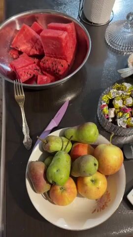 The video shows a close-up shot of a dark-colored table with various fruits and candies. In the upper left corner, there is a metal bowl filled with cut pieces of watermelon. Next to the bowl, a fork rests on the table. A purple-handled knife is positioned in the center of the frame, just below the watermelon bowl. To the right of the knife, a bowl filled with wrapped candies is visible. In the lower portion of the frame, a white bowl contains a mix of apples and pears. The lighting suggests an indoor setting, possibly a kitchen or dining area.