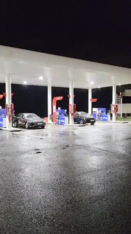 The scene is a gas station at night. The ground is wet, reflecting the light from the overhead canopy. The canopy is supported by white pillars and has red accents around the gas pumps. There are three cars parked at the pumps: a white van on the left, a gray sedan in the middle, and a dark-colored SUV on the right. A person is standing at the middle pump, presumably filling up their car. In the background, a white vehicle is on a trailer, and the sky is a dark, solid color, indicating nighttime.