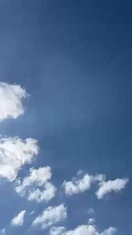 The video starts with a view of a blue sky with scattered white clouds. The camera then pans down to reveal the side of a building with a window and a roofline visible. The camera focuses on a black downhill mountain bike parked against the wall of the building. The bike has yellow accents on the handlebars, pedals, and rims. The ground around the bike is covered in grass and weeds.