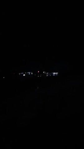 The video shows a very dark scene at night. In the distance, there are a few lights, likely from a building or town. The foreground is completely dark, and the audio suggests that the video was taken outdoors and captures the sound of wind.