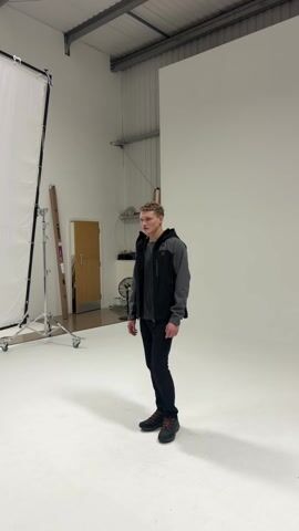 A young man stands in a white-walled studio. He wears black trousers, a gray t-shirt, a black and gray jacket, and black shoes with orange laces. He has short blonde hair. The studio contains a tall white screen on a rolling stand, a white backdrop, a door, a fan, and some miscellaneous objects. The man looks around.