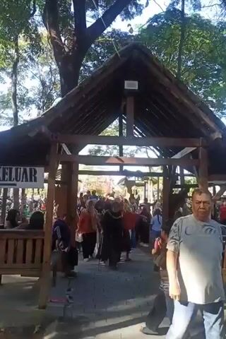 The video shows an outdoor scene with a wooden structure in the foreground. People are walking through an opening in this structure. The opening is covered by a roof with a triangular facade. To the left of the opening, there is a wooden bench and a sign that says "KELUAR," which means "EXIT" in Indonesian. The ground is paved with stone or brick. The background is blurred and appears to be a park or outdoor recreational area with trees and foliage. Sunlight is visible, suggesting it is daytime. The people are dressed in casual clothing, and some are wearing headscarves. They appear to be moving in the same direction, entering or exiting the area under the structure.