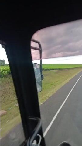 From inside a moving vehicle, the video pans across a vibrant green tea plantation under a cloudy sky. The camera angle shifts, revealing a winding asphalt road with a single yellow line marking the center, and a grassy shoulder on the left. A motorcyclist followed by a pedestrian walk in the distance along the road. The inside of the vehicle is seen briefly, showing the door frame and window.