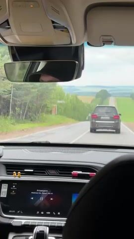 The video is filmed from the interior of a car, looking forward. The driver's seat is visible in the foreground on the right side. The dashboard has a large touchscreen display showing what appears to be a music player with Russian text. A gear shift is visible below the screen. Ahead, the road stretches into the distance, bordered by green fields and trees. A dark-colored SUV is driving in front, on the same road. The sky is overcast with some clouds. A rearview mirror is mounted on the windshield, reflecting a person wearing a dark baseball cap.