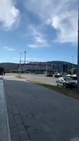 The video shows an urban street scene. The camera pans from left to right. It showcases a row of cars parked along the street, a shopping center in the background, a modern church with two towers, and apartment buildings. The sky is blue with scattered clouds. The street is paved with gray bricks, and there are trees and shrubs along the roadside. There are also trash receptacles.