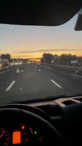 The video shows a view from the interior of a vehicle driving on a highway at dusk. The sky is a gradient of orange and gray, with the sun setting in the distance. The road ahead is visible, with lane markings and other vehicles, including a truck with its taillights illuminated. Trees and foliage line the sides of the highway. The windshield of the vehicle has several specks and smudges on it, which are visible throughout the video. The interior of the vehicle is dimly lit, with the dashboard lights glowing orange. The camera appears to be mounted inside the car, facing forward.