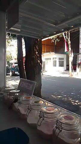 A medium shot captures a view through a window of several glass jars filled with what appears to be spices or toppings. Beyond the window, a tree casts shadows on a street. A black car drives past, followed by a person on a motorcycle. In the background, a white building stands with an Indonesian flag displayed. The scene is illuminated by daylight.