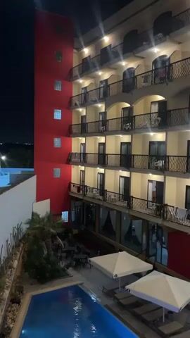 The video shows a multi-story hotel at night. The hotel has a red and tan exterior, with multiple balconies on each floor. Below, there is a pool surrounded by lounge chairs and white umbrellas. The pool is illuminated by lights. There are no visible people.