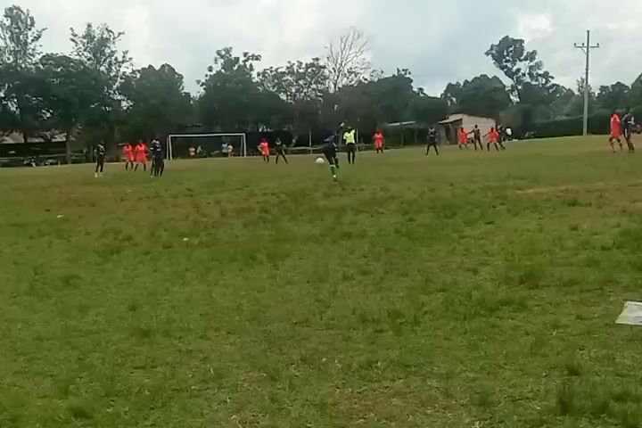 A wide shot shows a soccer game in progress on a grass field. Players wearing orange jerseys compete against players in dark blue jerseys. The soccer ball is in the middle of the field, surrounded by players. A soccer goal is visible in the background. Trees surround the field. Overhead, the sky is cloudy.