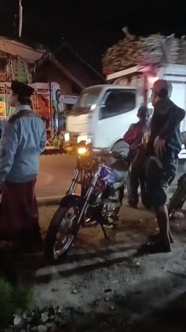 The video shows a nighttime street scene. In the foreground, a blue motorcycle is parked. To the left of the motorcycle, a person wearing a light blue shirt and a red sarong is standing, facing away from the camera. Behind this person, another motorcycle is visible. To the right of the blue motorcycle, several people are standing. One person, wearing a dark hooded sweatshirt and shorts, is looking down. Another person, wearing a maroon jacket and a cap, is looking towards the left. In the background, a large white truck with a cargo bed filled with what appears to be sugarcane is parked. The truck has red and white flashing lights on its rear. The environment appears to be a road with some debris on the ground. Buildings with pitched roofs are visible in the background, along with streetlights. There is a slight blur in the background, suggesting movement.