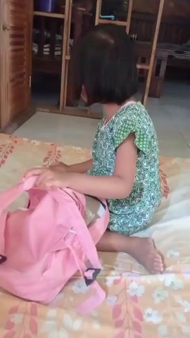 A young girl with dark hair and wearing a green and white patterned dress is sitting on a beige and pink floral patterned mat. She is in the process of interacting with a pink backpack. The girl is kneeling and her legs are bent. She has her hands on the top straps of the backpack. In the background, there is a wooden cabinet with shelves containing various items, including what appears to be a blue container. The lighting suggests an indoor setting. The girl's facial expression is focused on the backpack.