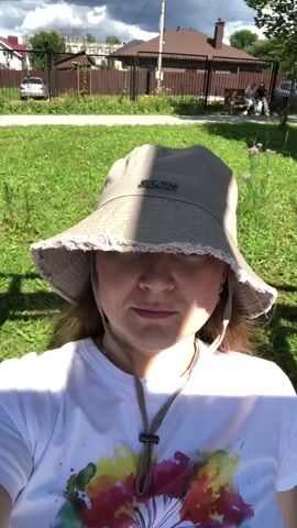 A person wearing a light-colored bucket hat and a white t-shirt with a colorful abstract design on the front is shown in a selfie video. The person is outdoors, with a background of green grass, trees, houses, and a fence. The sky is partly cloudy. The person blinks their eyes and slightly moves their head. There are other people visible in the distance sitting on a bench.
