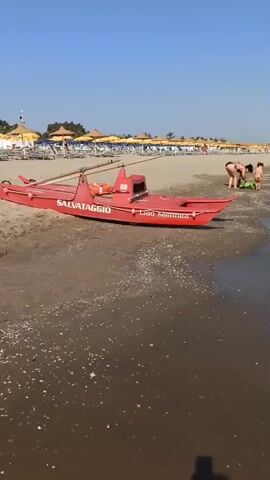 A red and white lifeboat is pulled up on a sandy beach. There are beach chairs and umbrellas set up behind the lifeboat. In the distance, people are visible on the beach. A plume of black smoke is rising into the clear blue sky above a line of trees and umbrellas.