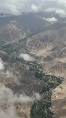 This aerial video showcases a vast, mountainous landscape in Tanglakpurna, India. The scene is dominated by rugged, arid mountains with subtle hints of green vegetation concentrated along a winding valley. Wispy clouds drift through the sky, partially obscuring the peaks in the distance which are capped with snow. The valley floor is lush with green fields, indicating agricultural activity and settlements within the otherwise desolate terrain. A road can be seen snaking through the valley, connecting the scattered villages. The overall impression is one of remoteness and natural beauty, with the juxtaposition of barren mountains and the vibrant green of the valley creating a striking visual. The time of day appears to be daytime, with diffused light suggesting either early morning or late afternoon, or possibly overcast conditions due to the clouds. No people or specific objects are clearly discernible due to the altitude. The atmosphere is serene and awe-inspiring, highlighting the grandeur of the natural environment. There is no visible text in the video.