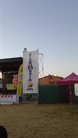 The video shows an outdoor event with a stage set up. A large white banner with the words "BIRRARDIA" and "AVIS" is prominently displayed, supported by scaffolding. To the left of the banner, a stage is visible, equipped with lighting and sound equipment. In front of the stage, a barricade separates it from the audience area. Several people are seated at tables in the foreground, some of whom appear to be eating or drinking. The environment is grassy, and the sky is clear, suggesting it is daytime or early evening. To the right of the stage, a green tent-like structure is partially visible. The overall scene suggests a festival or an outdoor concert.