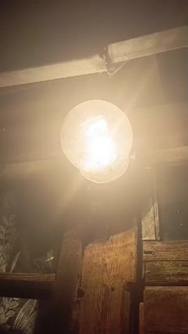 A close-up shot focuses on a lit incandescent light bulb hanging from a rough wooden beam. The bulb is illuminated with a warm yellow glow, casting shadows on the surrounding wooden structure and a patterned fabric visible in the background. The overall scene suggests an indoor, perhaps rustic or dimly lit environment. The camera remains stationary throughout the brief clip.