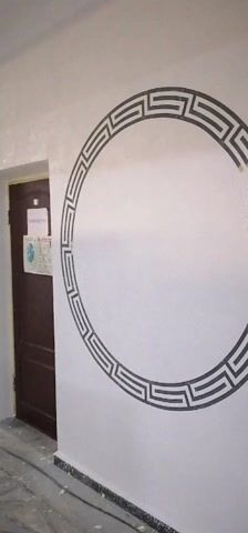The video pans along a corridor in a building. The walls are white with painted black designs. One wall has a large circle with a pattern reminiscent of a Greek key design. Other walls have rectangle designs with vertical lines and a diagonal line through the center. There are dark brown doors set into the walls. Two men walk down the corridor towards the camera. The floor is tiled. There is a metal frame on wheels toward the end of the corridor. The ceiling is white with a single light fixture.