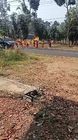 A group of people in red and yellow outfits are marching in a parade on a paved road, holding flags. There are several vehicles, including cars and motorcycles, present on the road. The environment appears to be rural, with trees and vegetation lining the road. The ground in the foreground is covered with dirt and debris.