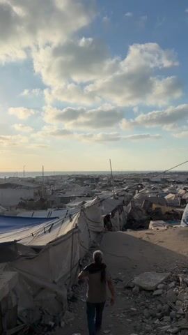 A person walks along a dirt path through a densely packed encampment of temporary shelters made of tarp and canvas. The shelters are arranged in rows, creating narrow walkways between them. In the background, the ocean is visible under a cloudy sky. The ground is uneven, with debris and rocks scattered around. The person is walking away from the camera, towards the encampment. The overall impression is one of a makeshift settlement.