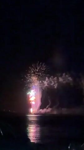 The video shows a nighttime fireworks display over water. Brightly colored fireworks, including bursts of yellow, green, blue, and pink, explode in the dark sky. A large plume of pink and white smoke descends from the fireworks, creating a waterfall-like effect over the water. The fireworks' reflections shimmer on the surface of the water, adding to the visual spectacle. The surrounding environment is dark, with the exception of the illuminated fireworks and their reflections.