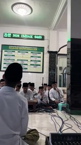 The video shows a group of young boys in white shirts and black caps sitting on the floor in front of a microphone and what appears to be a soundboard. They are in a room with tiled walls and a green organizational chart on the wall that says "STRUKTUR ORGANISASI PENGURUS TAKMIM MASJID BAITUSSALAM" which translates to "Organizational Structure of the Takmir Management of Baitussalam Mosque". A digital clock on the wall displays "21 IM5AK 04:03 SUBUH". There is a man on the right side of the frame also speaking into a microphone. The overall scene appears to be inside a mosque.