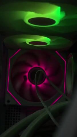 This is a close-up video of two computer case fans. The bottom fan is larger and glows with a bright pink light. It has two tubes running through its center. The top fan is smaller and is lit with a shifting combination of green, blue, and purple lights. The fans are mounted inside what appears to be a computer case, with some dark metallic structures visible in the background and to the right. The lighting in the video is dim, with the colorful glow from the fans being the primary light source. The video appears to be taken from a slightly upward angle, looking into the case.