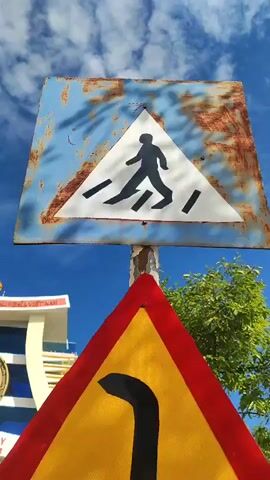 This is a vertical, outdoor, medium shot video of road signs with a border crossing gate in the background. The top sign is a weathered, blue metal square with a white triangle in the center showing a black pedestrian silhouette walking on a crosswalk. The bottom sign is a yellow triangle with a red border, displaying a black curved arrow pointing to the left. A white pole supports both signs. In the background, a modern, beige and white gate stands against a bright blue sky with scattered clouds. The gate has arches and a large, circular emblem with text in Vietnamese and English that reads "BO Y INTERNATIONAL BORDER GATE". A Vietnamese flag is visible to the right of the gate. The ground in front of the gate appears to be a paved road, and vegetation and trees are visible on either side. The overall scene suggests an international border crossing in Vietnam.