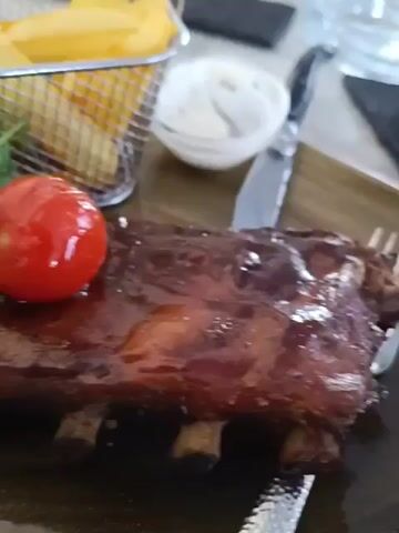 The video shows a close-up view of a plate of food, with a rack of ribs in the foreground. The ribs are glazed and appear to be barbecued. They are garnished with a cherry tomato and some green sprouts. To the left of the ribs, there is a small bowl of red sauce, and behind that, a basket of french fries. In the background, a white dip is visible, along with silverware. The table surface appears to be wooden. The lighting is bright, and the focus shifts slightly throughout the short clip.