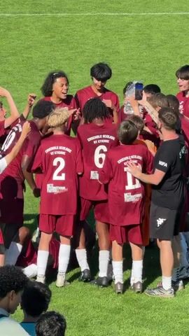 A group of about a dozen adolescent soccer players, wearing matching maroon uniforms, celebrate on a sunny grass field. They are tightly huddled together, with some players having their arms around each other. The uniforms read "ENSEMBLE SCOLAIRE ST ETIENNE" and include jersey numbers.  One player holds a large plastic water bottle over the group and pours water over their heads. A man in a black "STAFF" t-shirt and black shorts stands among the players, holding them back slightly with his hand. One person appears to be recording the celebration with a smartphone.  The players are laughing and jubilant. The scene suggests they have just won a game or achieved a victory.  Another person is also partially visible kneeling in the foreground, outside of the huddle.