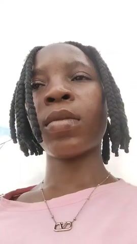 The video is a close-up, low-angle shot of a young Black person with dark braided hair. The person is wearing a pink t-shirt and a silver necklace with a pendant. The background appears to be an outdoor setting with a cloudy sky. The person's face is the primary focus, and their gaze shifts slightly throughout the short clips. There are no other people or significant objects visible in detail.