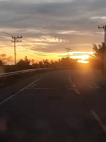 The video captures a first-person perspective from a moving vehicle on a road during sunrise or sunset. The sun, a bright orange orb, is visible at the horizon, casting a warm glow and creating lens flare. The road is dark asphalt with a white solid line and a yellow broken line. On the left side of the road, there's a guardrail and some sparse vegetation. On the right, there are telephone poles with multiple wires and a dense line of trees. The sky above is cloudy, with shades of grey and hints of orange near the horizon. The vehicle appears to be moving on a slight curve to the right.