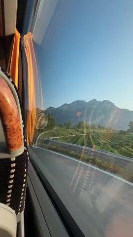 The video is filmed from the perspective of a passenger inside a moving vehicle, looking out a window. The window has an orange curtain on the left side. Outside the window, a road with a metal barrier runs alongside a field of green crops, possibly vineyards, which are laid out in neat rows. Beyond the field, there are trees and a large, rocky mountain face under a clear blue sky. The sun is casting a warm light on the landscape, creating reflections on the window. The vehicle appears to be traveling along a highway or a similar paved road. The perspective shifts slightly as the vehicle moves, causing the landscape to slide by. The video ends as the vehicle approaches a tunnel entrance.
