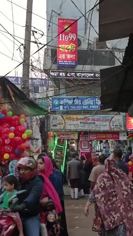 The video shows a crowded street in what appears to be a market area in Bangladesh. Many people are walking through a narrow lane lined with shops and businesses. Signs are visible above the shops, written in both English and Bengali. Overhead, a tangle of wires is visible. A vendor stands on the left selling bunches of balloons. The people are dressed in a variety of traditional and modern clothing. The atmosphere is lively with activity.